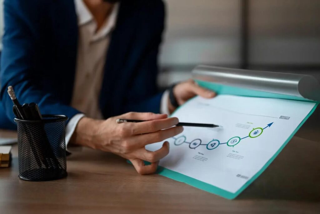 Na zdjęciu znajduje się osoba w garniturze, która trzyma dokument zawierający diagram. Osoba wskazuje na ten diagram długopisem, co sugeruje, że omawia przedstawiony plan lub proces.