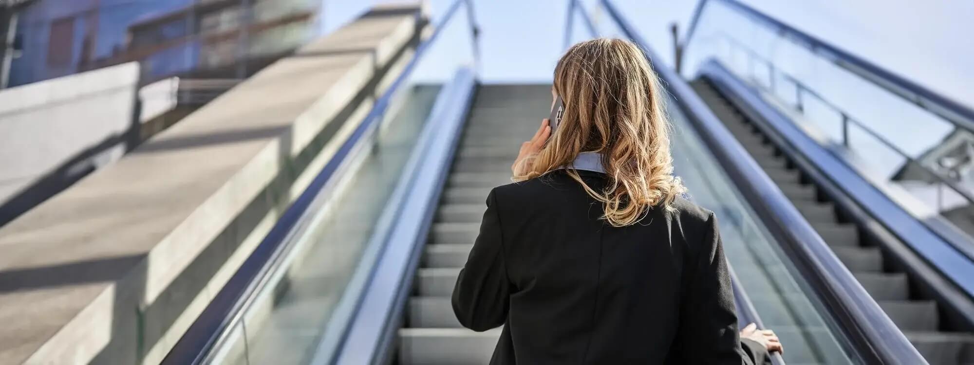 Na zdjęciu kobieta ustawiona do kamery tyłem. Kobieta ma blond włosy do ramion, ubrana jest w czarną marynarkę i niebieską koszulę. Stoi na zewnątrz, na ruchomych schodach i trzyma telefon przy uchu.