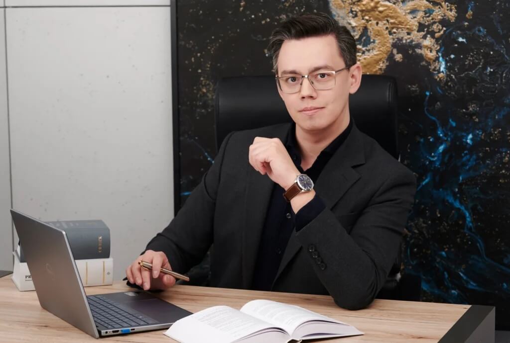 Na zdjęciu Mateusz Haśkiewicz ubrany w czarny garnitur i czarną koszulę oraz nosi zegarek na ręce. Mężczyzna siedzi przy drewnianym biurku. Przed nim znajduje się laptop, otwarta książka oraz długopis trzymany w dłoni. W tle widzimy ciemną ścianę ozdobioną kolorowym, abstrakcyjnym wzorem.